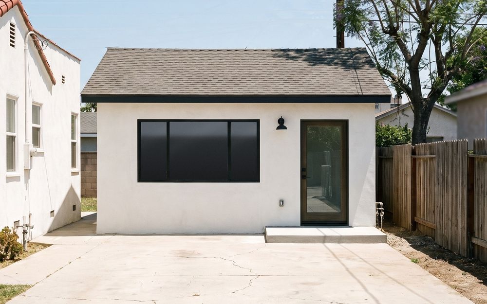 A detached two-car garage turned into a small home, seen straight on from the end of a cracked concrete driveway, with a wide black-framed window and a glass entry door where the roll-up door was